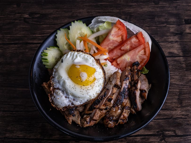 Grilled chicken, fried egg, rice, and fresh vegetables in a black bowl on a wooden table.