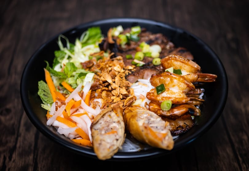 A black bowl of vermicelli noodles with grilled shrimp, pork, spring rolls, and vegetables.