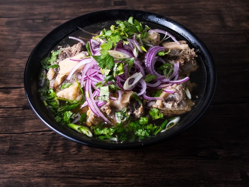 A black bowl of oxtail pho with red onions and cilantro on a dark wooden table.