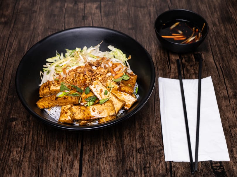 A black bowl of vermicelli noodles with tofu, vegetables, and sauce, with chopsticks and dipping sauce.